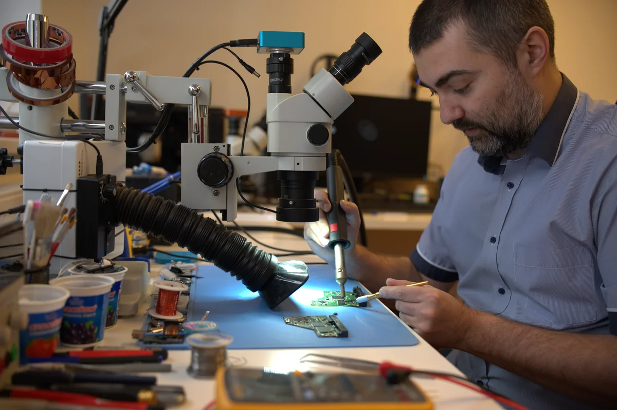 Senior technik ITHOPE páje pod stereomikroskopem PCB; pracovní stanice s odsávačem výparů, měděná cívka a multimetr
